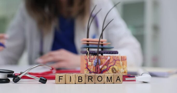 Wooden blocks spell word Fibroma beside skin model on table. Woman dermatologist studies patient medical case writing detailed therapy notes