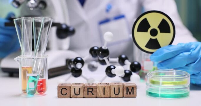 Wooden cubes on lab bench spell word Curium near international radiation symbol. Scientist in gloves prepares sample test observing atomic model