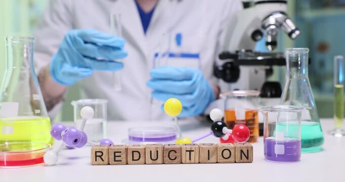 Wooden blocks on desk spell word Reduction near vivid chemical samples in glassware. Chemist transfers reagent with pipette studying redox change