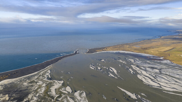 Aerial view of glacial river mouth meeting the dark ocean, creating a stark contrast of colors and textures under a vast sky, Iceland, Iceland.