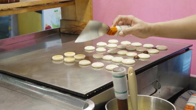 Close-up of a street food vendor placing batter into a circular iron pan to make sweet coconut desserts in Bangkok.