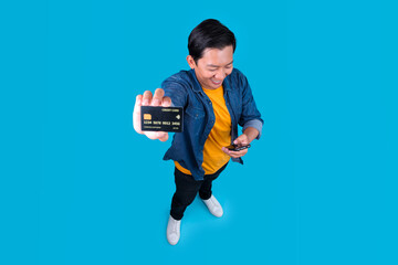 Asian Man Looking At Smartphone While Showing Credit Card Ready For Mobile Banking on Blue Background