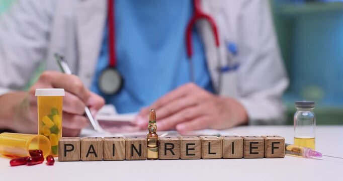 Doctor reviews symptoms and writes preliminary diagnosis. Wooden cubes arranged on desk form phrase Pain Relief symbolizing purpose of consultation