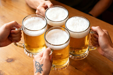 Four hands clinking frosty mugs of golden lager on a wooden table, celebrating together