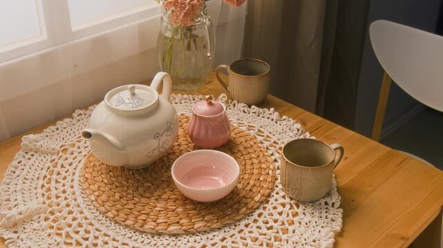 Medium close up shot of ceramic teapot, cups, sugar jar and bowl on woven placemat over lace doily on wooden home table
