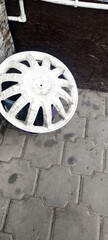 A dirty, white plastic wheel cover lies on a grey interlocking paved surface, resting against a dark wooden background and white pipes