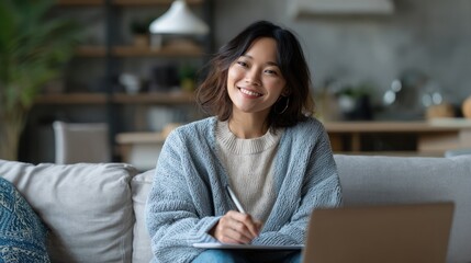 Smiling Woman Writing Notes on Gray Sofa at Home with Laptop