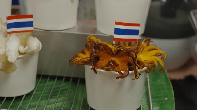 Close-up of fresh seafood skewers including grilled squid, garnished with flowers and small Thailand flags at SookSiam indoor market.