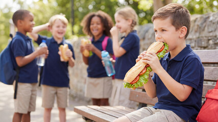 Group of five school-age children in navy polos and khaki pants eat sandwiches outdoors on sunny day. A cheerful moment of lunch, friendship, and healthy habits in a natural schoolyard setting.