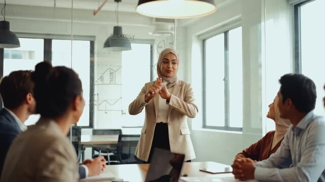 Muslim businesswoman leading a strategy presentation in a modern office. Female leader in a hijab explaining charts on a glass board to a diverse team. Corporate meeting and collaboration concept