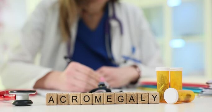 Wooden blocks on medical desk spell word Acromegaly beside pills containers. Woman doctor writes notes about growth disorder studying patient chart