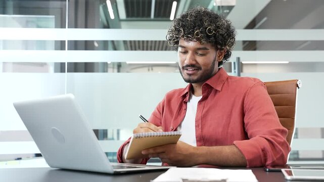 Young businessman watching video call conference or training notes in notebook looking at laptop screen sitting at workplace in business office. Manager communicates remotely at an online meeting