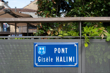 The scene captures the Gisele Halimi Bridge in Mont de Marsan. The striking blue sign is surrounded by vibrant greenery. Quiet streets reflect a peaceful atmosphere, inviting exploration.