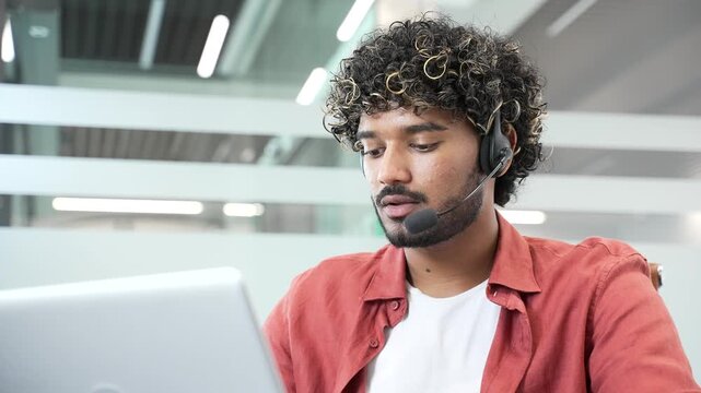 Young smiling male corporate operator in headset working in customer support service on helpline telesale talking using laptop in business office. Agent of call center advises client online. Close up