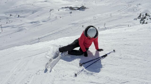 Woman skier in red jacket getting up from snow after falling down on steep ski slope at crowded mountain resort on sunny winter day. Leg and body injuries from a fall on a ski piste