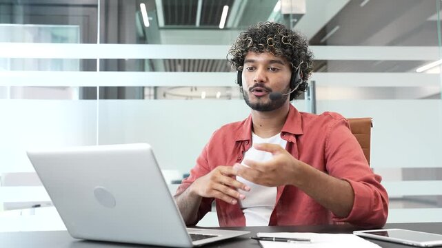 Young handsome businessman in a headset talking on a video call using laptop computer sitting at workplace in office. Entrepreneur has a business meeting. Manager speaks remotely at online conference