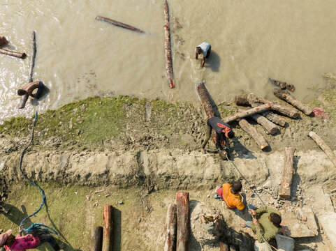 Bagerhat, Bangladesh - 05 December 2025: Aerial view of workers struggling to move logs in the muddy river, their vivid clothes contrasting with the dull landscape.