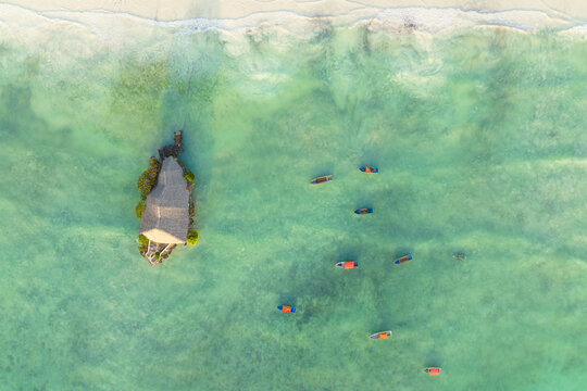 Aerial view of vibrant turquoise water gently laps against a small island with a thatched-roof structure, surrounded by local boats, Zanzibar, Unguja South Region, Tanzania.