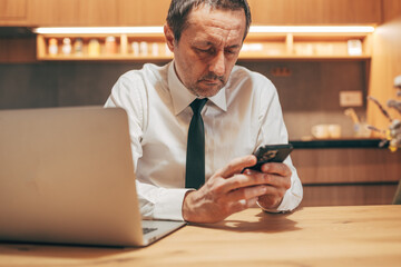 Mature professional using laptop and mobile phone simultaneously, managing work responsibilities in a modern home office environment.