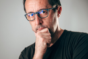 Serious middle-aged man wearing glasses, hand on chin, looking at camera with thoughtful expression, symbolizing intelligence, analysis, and critical thinking.