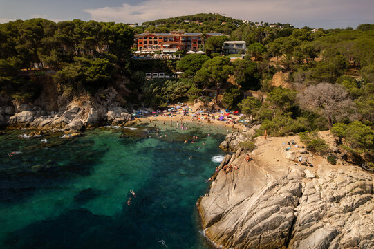Aerial view of azure waters meet golden sands at Cala del Pi, where sunbathers bask below the hotel's terracotta facade, Platja d'Aro, Catalonia, Spain.