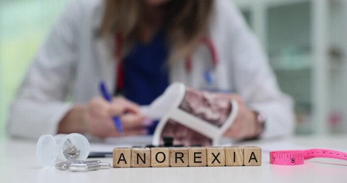 Wooden blocks spell word Anorexia on medical desk in clinic. Woman doctor studies patient scan pictures while noting weight loss disorder carefully