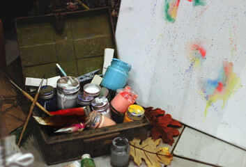 Vintage art supplies in an old wooden box: colorful jars of paint, brushes, and a dry leaf next to a messy canvas, symbolizing creative process and artistic inspiration