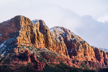 Rocky, rugged red sandstone hillside in Sedona, Arizona. Top is sprinkled with snow, cloudy sky is in the background. Sun is shining on one side; green of the Coconino forest is at the base. 
