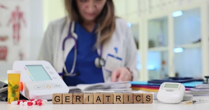 Wooden blocks on medical desk spell word Geriatrics near pressure monitor. Woman doctor reviews elderly patient chart planning long term care strategy