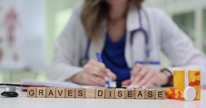 Wooden blocks spell words Graves Disease on medical table near pill containers. Woman doctor writes notes adjusting stethoscope and planning therapy