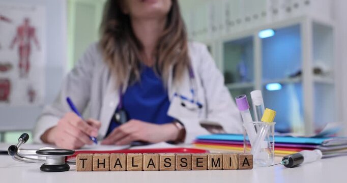 Wooden blocks spell word Thalassemia on lab desk near test tubes and stethoscope. Woman doctor writes hematology notes reviewing anemia management