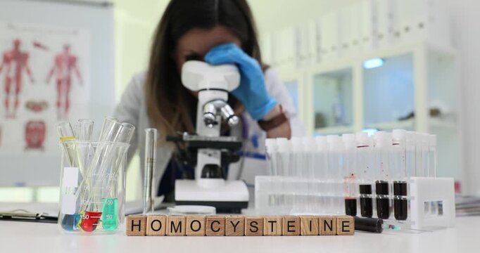 Wooden blocks on lab bench spell word Homocysteine at test tubes. Woman scientist looks through microscope studying blood plasma sample in laboratory