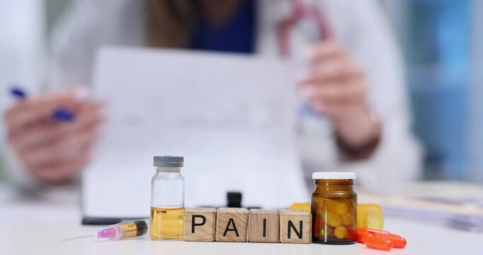 Wooden blocks on desk spell word Pain beside vials and syringe. Woman doctor reads chart writing detailed note about persistent pain control plan