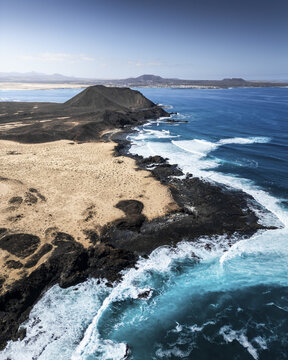 Aerial view of turquoise waves crashing against the black volcanic coast, meeting the arid landscape, Isla de Lobos, Canary Islands, Spain.