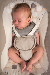Young infant wearing a striped bodysuit rests peacefully in a baby bouncer. The baby is supported by a soft harness and the patterned seat allows for comfortable sleeping.