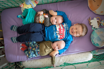 An older boy is smiling while lying in a crib. He has two younger baby brothers lying on top of him. All three kids are wearing clothes, laying on a purple surface.