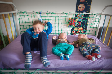 A young boy, wearing a blue shirt and pants, lays in a crib with his infant twin siblings. The twins are lying down, while the older brother is looking at the camera.