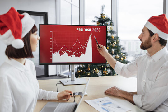 Fototapeta Close up of festive businessman manager in Santa hat sitting near big digital wall screen with company financial and growth charts and talks to multiethnic colleagues discussing results of project.