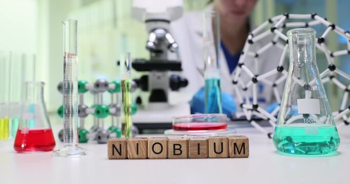Woman chemist examines Niobium and takes notes in paper notebook by microscope. Wooden cubes form word Niobium on table near test tubes with solutions