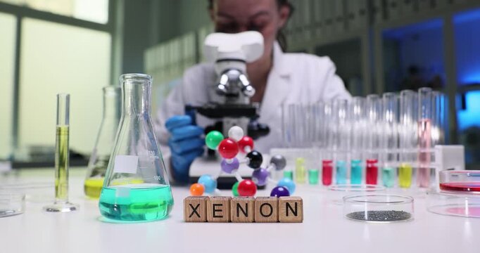 Woman chemist leans over microscope to examine tiny details of sample. Wooden cubes forming word Xenon serve as reminder of research context in lab