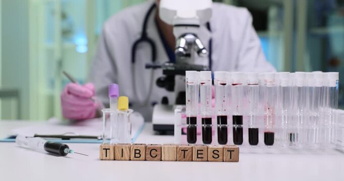 Woman doctor observes viscosity and color of blood at microscope and takes meticulous notes in notebook. Wooden cubes form phrase Tibc Test on table