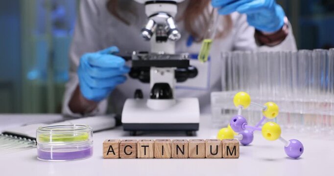 Woman chemist leans over microscope and holds test tube filled with bright yellow liquid near glassware. Wooden cubes form word Actinium on table