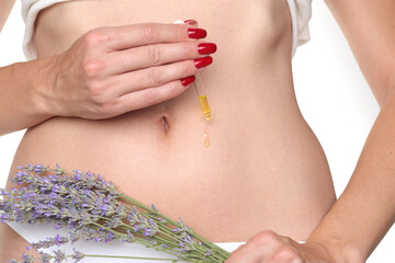 Close-up of a woman applying cosmetic oil with a dropper on her belly near the navel while holding lavender flowers, studio shot on a white background.