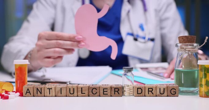 Doctor holds neatly cut paper model shaped like human stomach. Wooden cubes form phrase Antiulcer Drug hinting at recommended course of treatment