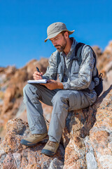 Man in outdoor gear sits on rocky terrain, writing in a notebook, surrounded by natural landscape, showcasing exploration and creativity in nature
