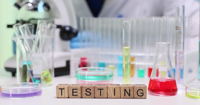 Woman laboratory worker leans slightly over microscope to study sample. Little wooden cubes arranged neatly on table form word Testing at glassware