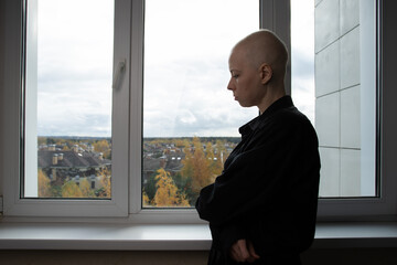 Portrait of a bald woman in black clothing standing by a bright window, gazing at the city view. The soft light and autumn colors,recovery after illness, cancer awareness, and mental resilience