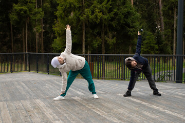 Two women practicing yoga outdoors in a forest park, one of them a cancer survivor wearing a headscarf and warm sportswear. They perform stretching and balance poses together