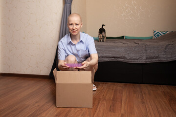 Bald woman after chemotherapy smiling while unpacking a parcel and holding a pink item, concept of recovery, positive emotions, and home comfort.