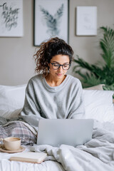 Young woman with curly hair, wearing cozy sweater and glasses, is working on laptop in a stylish bedroom with plants and artwork, creating a serene home office atmosphere